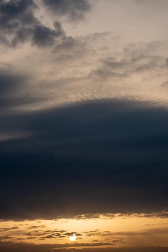 A cloudy sky during sunset, a large dark cloud comes across the middle, the sun can be seen at the bottom