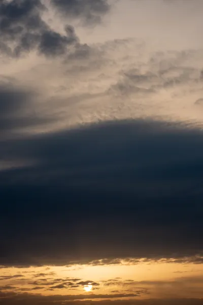 A cloudy sky during sunset, a large dark cloud comes across the middle, the sun can be seen at the bottom