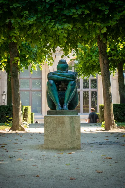In a small square, with trees all around, a bronze statue of a woman, sitting down, head in its arms