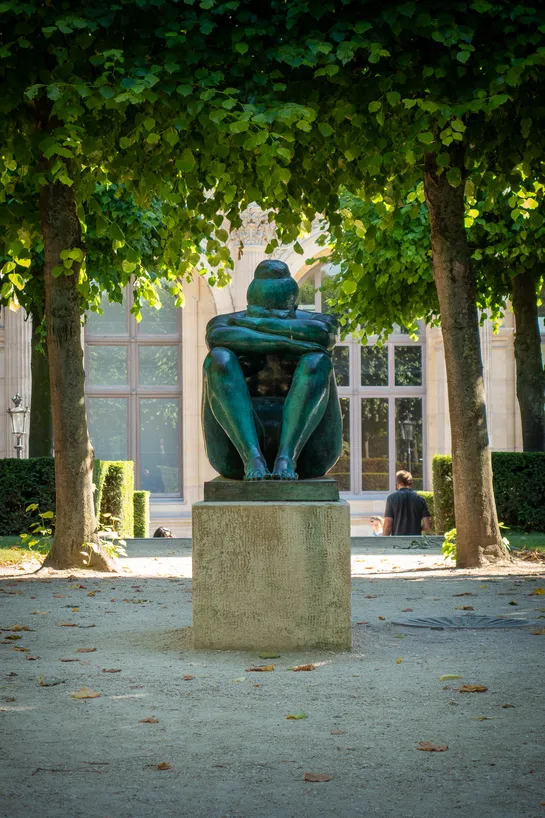 In a small square, with trees all around, a bronze statue of a woman, sitting down, head in its arms
