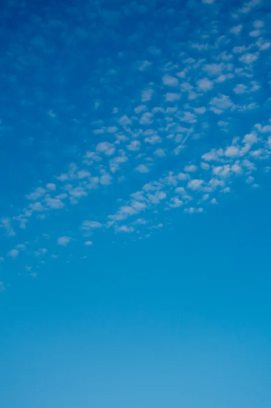 A deep blue sky, some clouds dotting the top, behind them a plane very far away leaving a trail