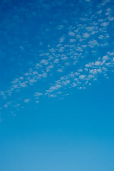 A deep blue sky, some clouds dotting the top, behind them a plane very far away leaving a trail