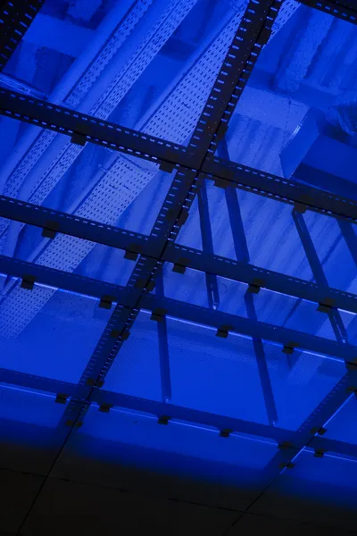 The ceiling of a large room, lit in blue, with multiple pipes and cable holders, all protected by a sort of metal grid