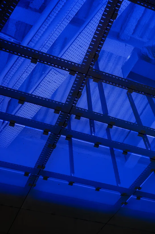 The ceiling of a large room, lit in blue, with multiple pipes and cable holders, all protected by a sort of metal grid