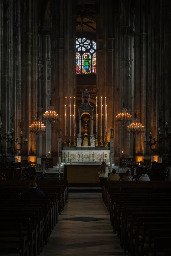 A church interior, dimly lit. Surrounded by very tall columns, few people on the pew and in the center, an altar with a lot of candles. At the very top, stained glass letting in a bit of exterior light.
