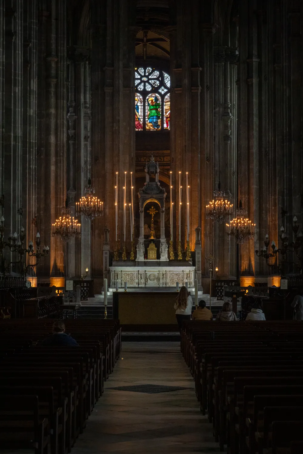A church interior, dimly lit. Surrounded by very tall columns, few people on the pew and in the center, an altar with a lot of candles. At the very top, stained glass letting in a bit of exterior light.