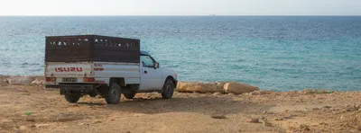 A pickup truck parked on top of a cliff, facing the sea. Far away on the horizon, the silouette of a ship