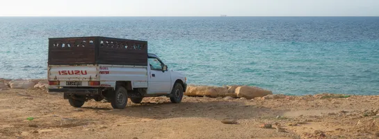 A pickup truck parked on top of a cliff, facing the sea. Far away on the horizon, the silouette of a ship