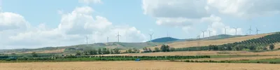 Wind turbines in the background, atop countryside hills, crops as far as the eye can see, a lone blue car speeds through in the middle