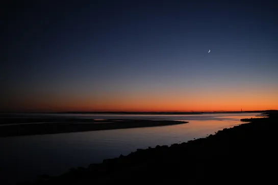 The sea shore, after the sun has set, the sky is a gradient of red to blue, and a moon crescent