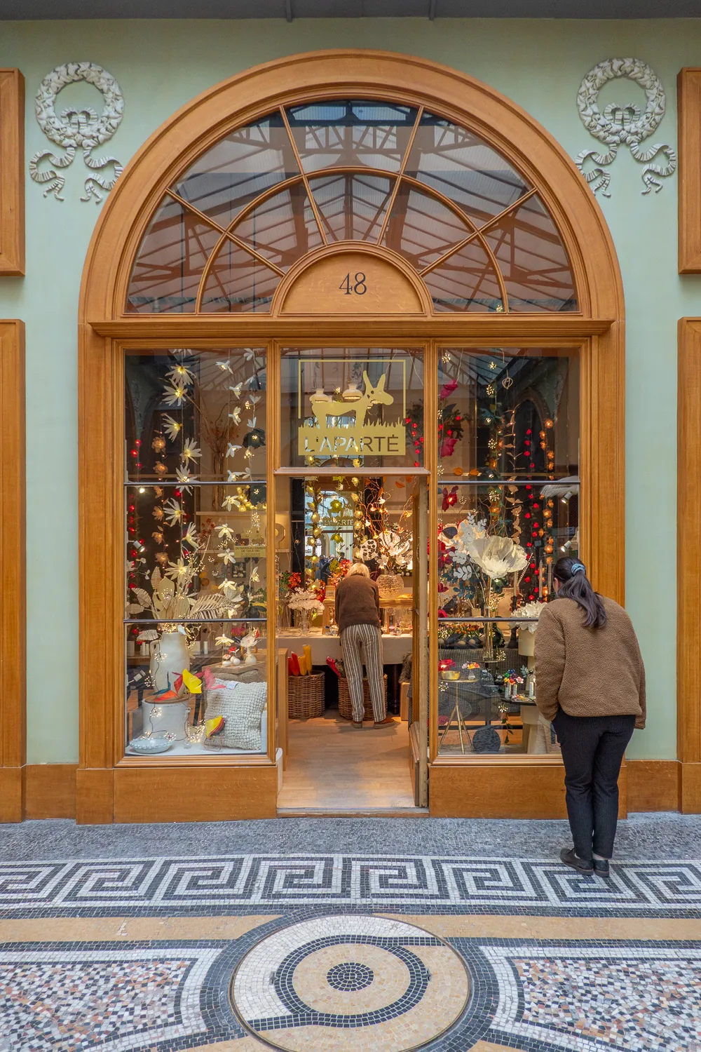 A quaint store front. One person on the right looking at wares, through the open door an employee tending to the shelves.