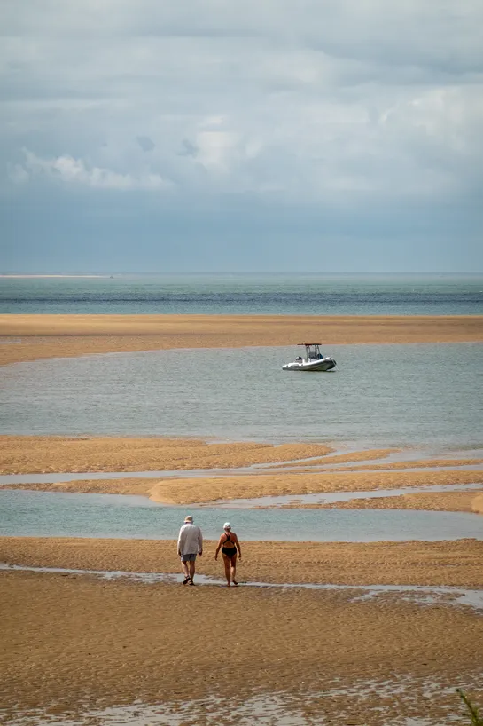 A small boat is anchored on a small bay formed by a sand bank, with two people walking on it