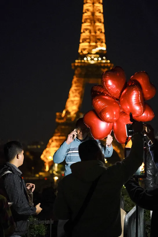 A person taking a phone call while holding red heart ballons, being lit by a spotlight, standing in front of the Eiffel Tower in the distance