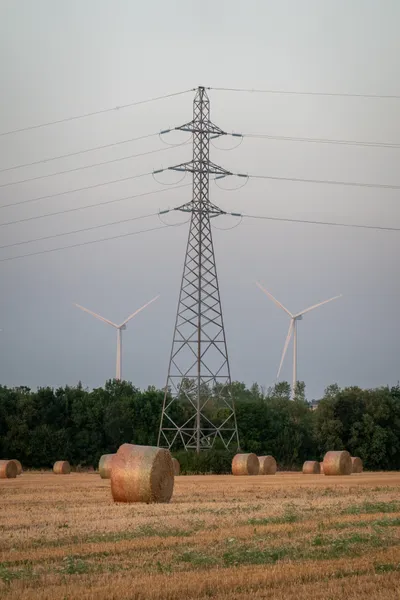A transmission tower in the center, in the background are two wind  a farming field in fron