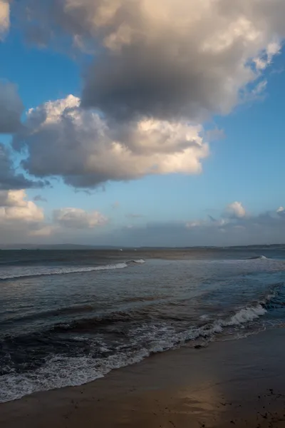 The beach, dimly lit. Calm sea. Large clouds above, the white reflected by the sea and the wet sand.