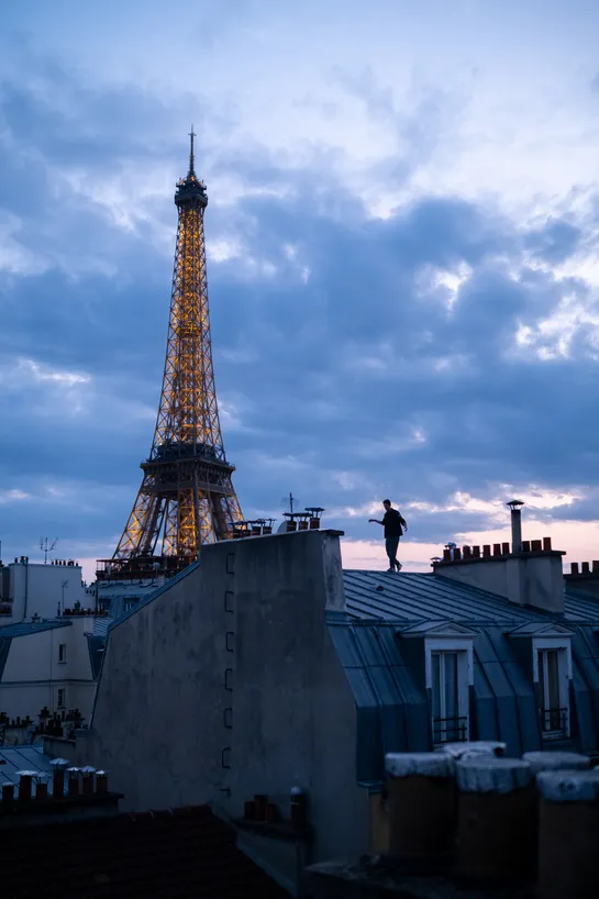 As the Eiffel Tower is visible on the left, the bottom of the frame is occupied by metal rooftops and chimneys typical of Paris ; a man walks in equilibrium on a rooftop