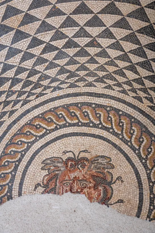 A roman mosaic representing Medusa, the bottom has been lost, we can only see part of her face. Above are decorative triangles forming concentric circles.