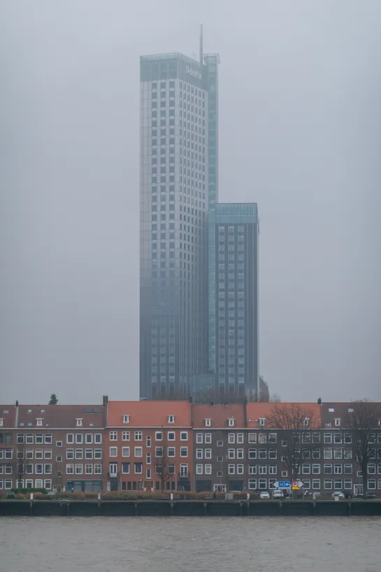 Grey sky, a tall skyscraper in the center. In front is a row of houses with orange or brown rooftiles and bricks, they face a canal.