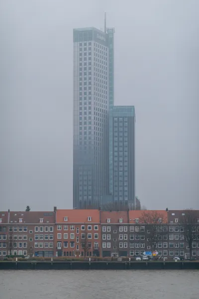 Grey sky, a tall skyscraper in the center. In front is a row of houses with orange or brown rooftiles and bricks, they face a canal.
