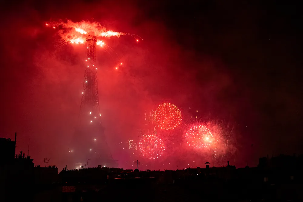 Red fireworks firing off, both from the grounds and the top of the Eiffel Tower, itself sparkling ; the red in the sky can be seen in reflections on the rooftops