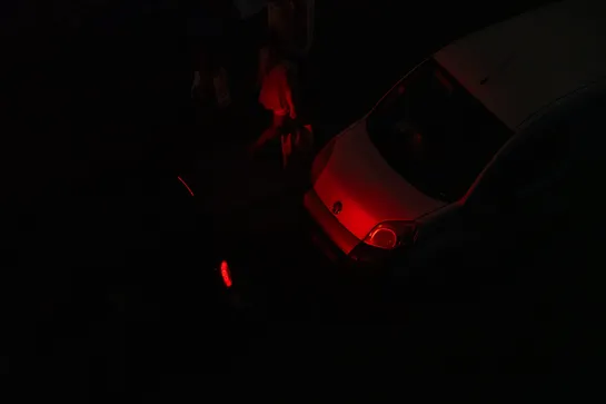 A car's red lights illuminates the car behind and passerbys