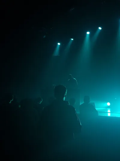 Amid the audience, one person looks at the artist playing music during a concert, almost no light except  blue spotlights, only etching the contours of people