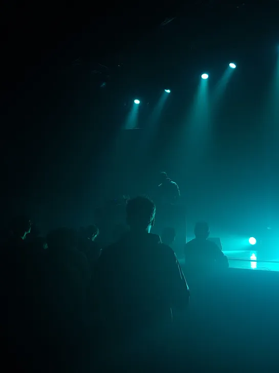 Amid the audience, one person looks at the artist playing music during a concert, almost no light except  blue spotlights, only etching the contours of people