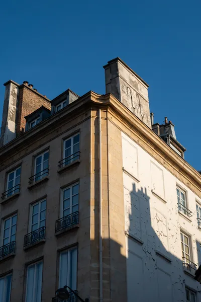 A building seen from its angle. The left part has windows in the shade. The right part is lit by the sun but there are no windows, the shadow of a chimney appears there as well.