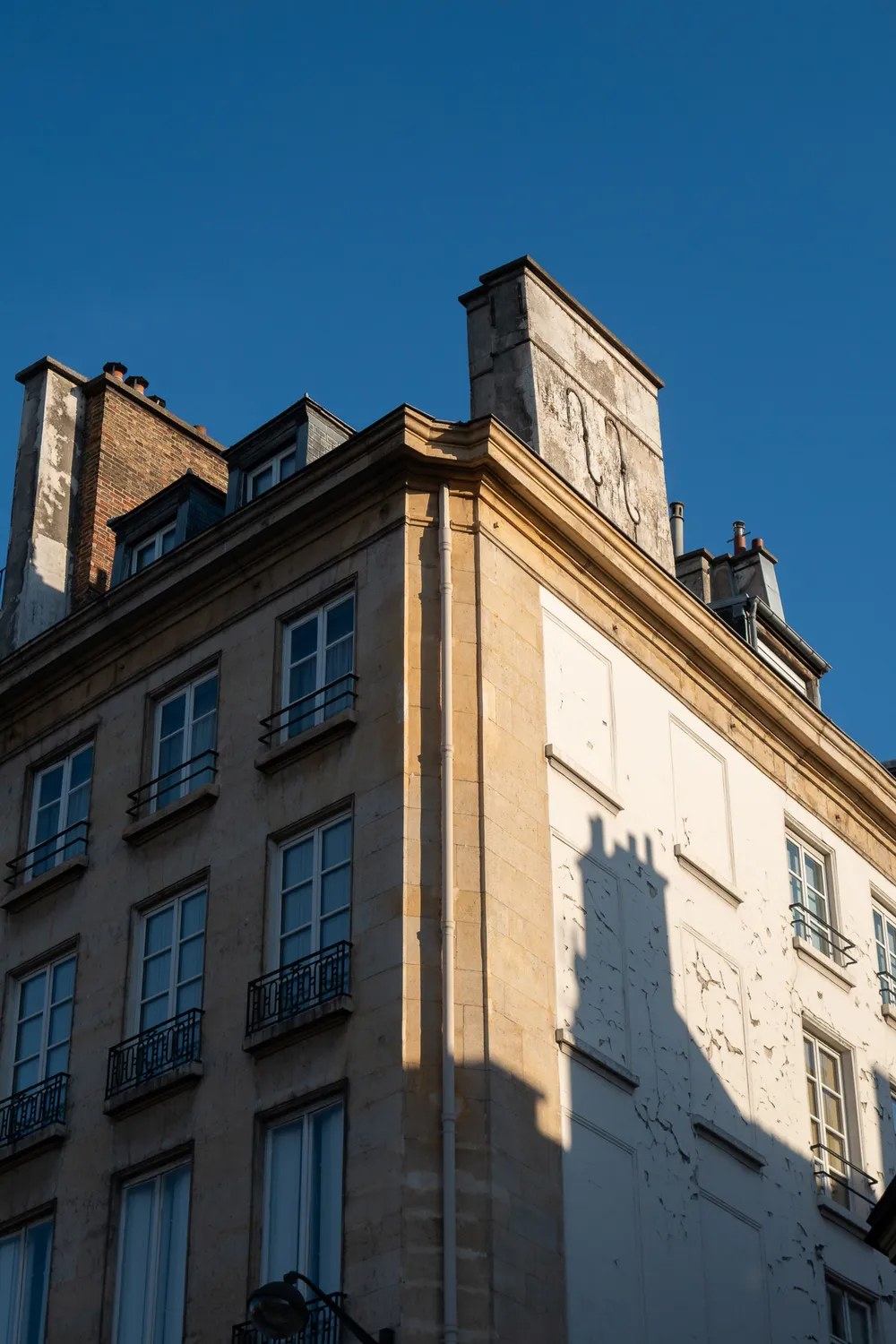 A building seen from its angle. The left part has windows in the shade. The right part is lit by the sun but there are no windows, the shadow of a chimney appears there as well.