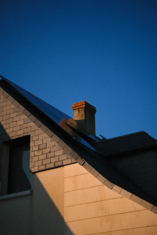 Beneath a blue sky, a rooftop with a chimney and solar panels, illuminated by the sunset