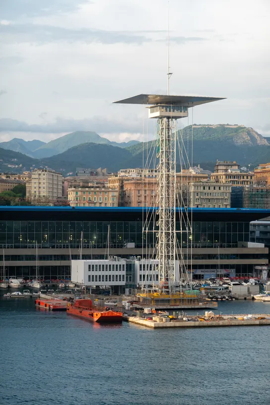 A large metal structure near the sea, city in the background, green mountains further away