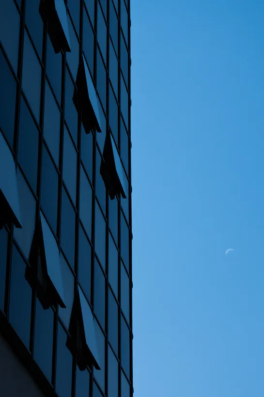 Modern building on the left reflecting the blue sky on the right, the moon is slightly visible in a crescent shape
