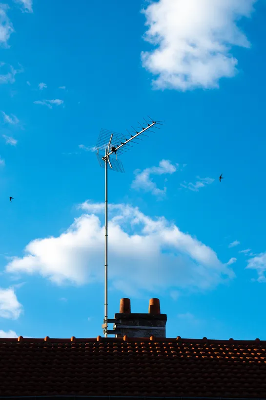 A clear blue sky with some clouds, in the foreground a house rooftop with a traditional antenna and a small chimney. Two small birds flying above.