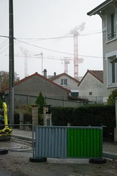 A city road during public works. Houses behind, not enough windows for all of them. Cranes can be seen in the background, somewhat visible through the fog.