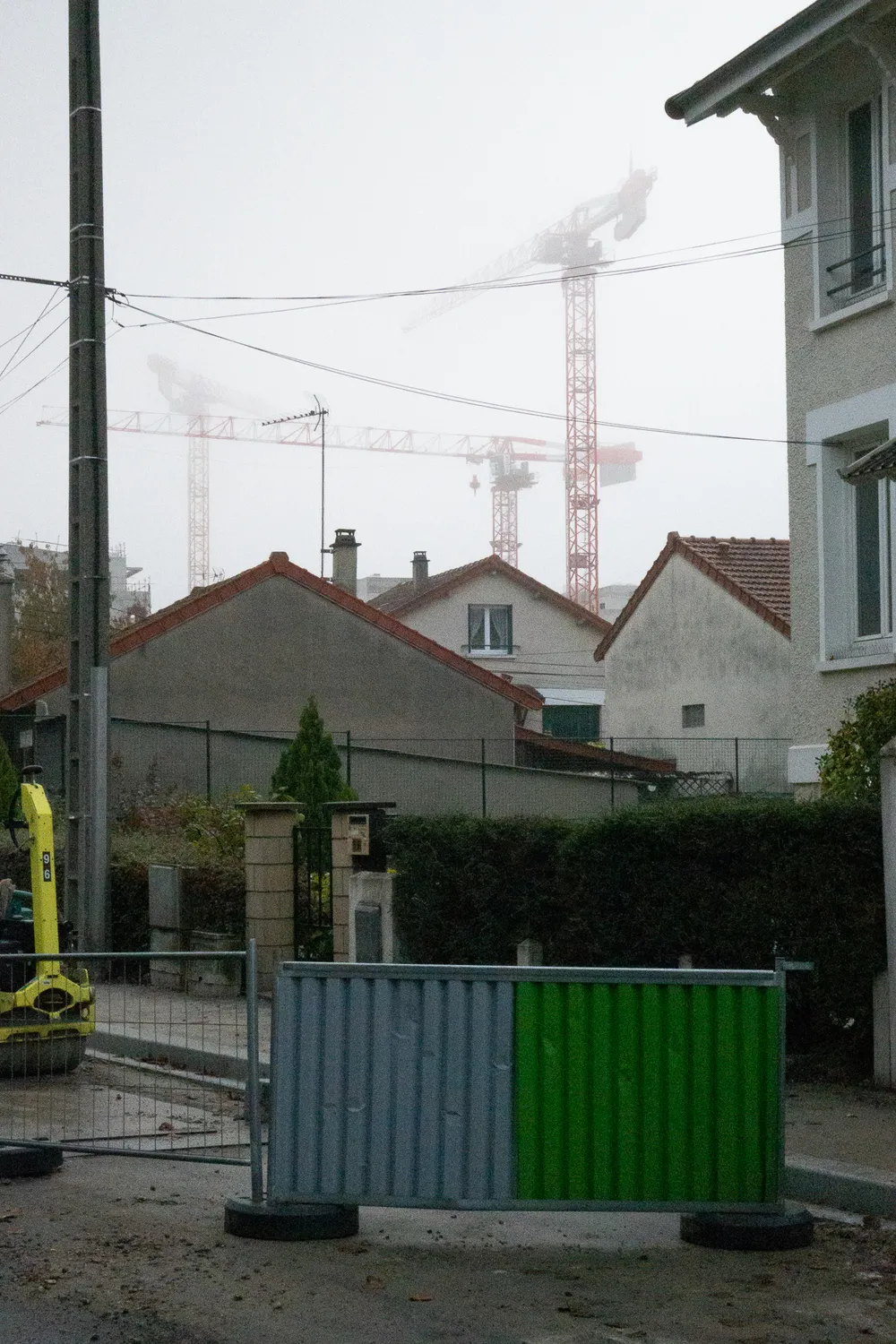 A city road during public works. Houses behind, not enough windows for all of them. Cranes can be seen in the background, somewhat visible through the fog.