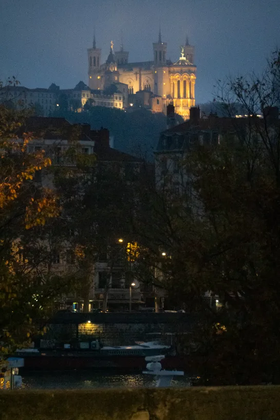 Nearly night, a tall church is lit in yellow from spotlights far away on the hill side. Nearer, few buildings, streetlamps, trees with autumn leaves, and a small portion of it all reflected on the river below