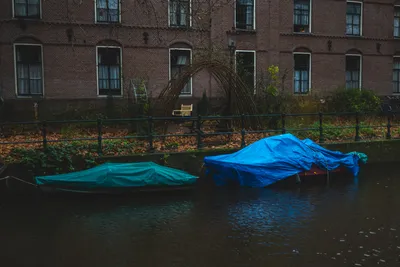 In front of a brick building, a water canal, two boats within, both covered with tarps, one green the other blue, both reflected in the water