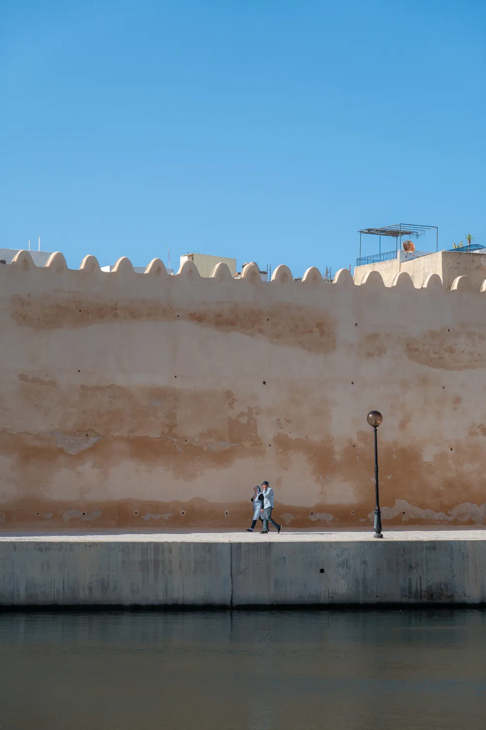 Two people walking side by side on a quay, alongside the medina wall, water beneath