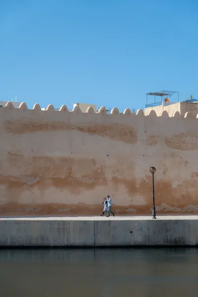 Two people walking side by side on a quay, alongside the medina wall, water beneath