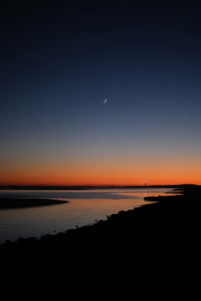 The sea shore, after the sun has set, the sky is a gradient of red to blue, and a moon crescent. In the distance, the lighthouse's beam is reflected on the waters.