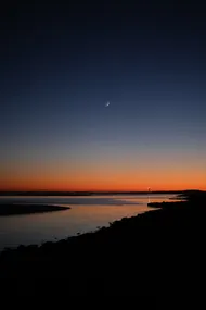 The sea shore, after the sun has set, the sky is a gradient of red to blue, and a moon crescent. In the distance, the lighthouse's beam is reflected on the waters.