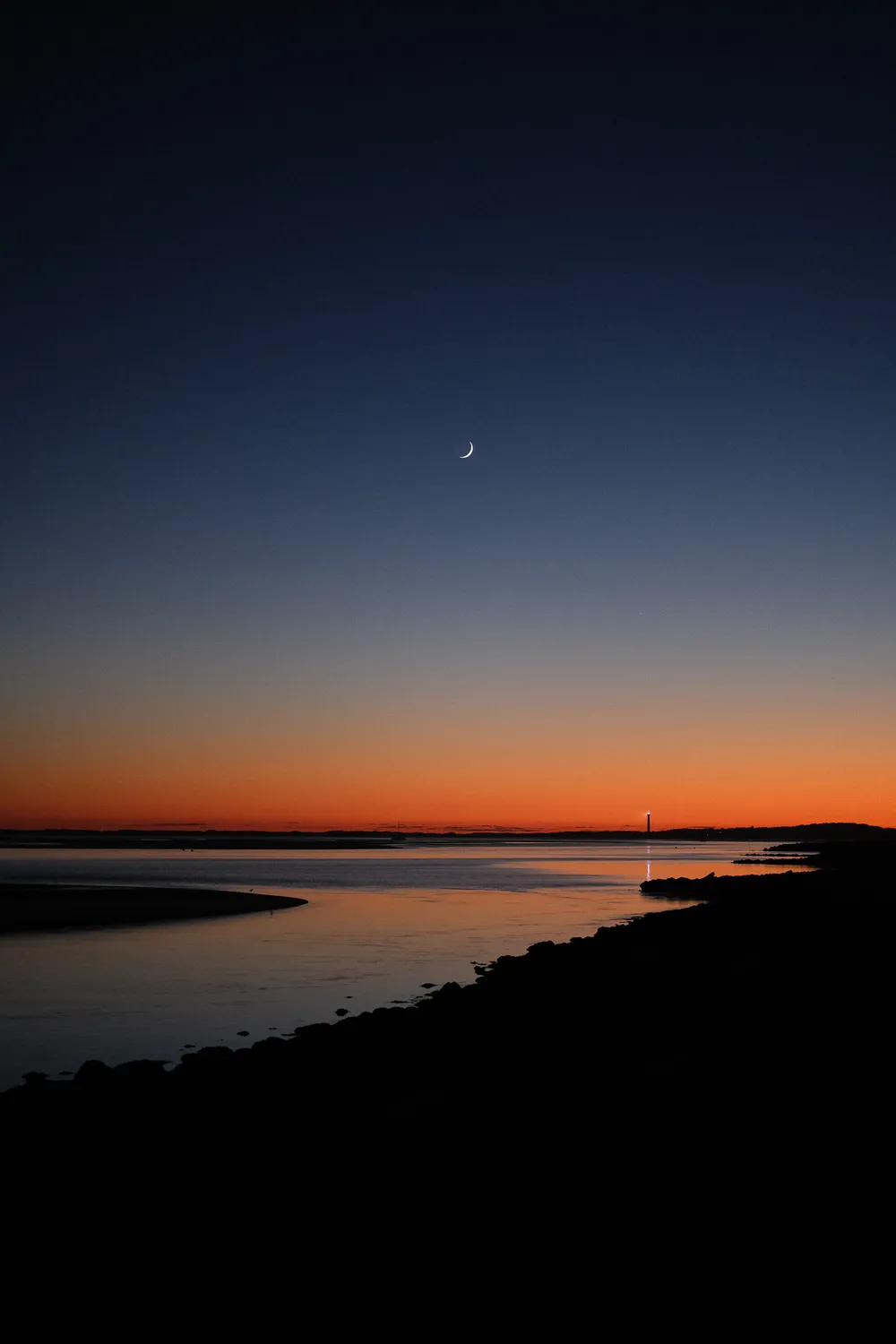 The sea shore, after the sun has set, the sky is a gradient of red to blue, and a moon crescent. In the distance, the lighthouse's beam is reflected on the waters.